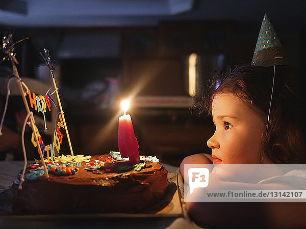Seitenansicht eines süßen Mädchens mit Blick auf Geburtstagskuchen auf dem Tisch in der Dunkelkammer