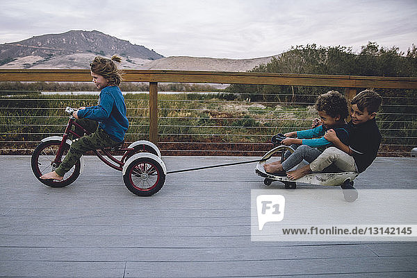 Junge zieht Spielzeugauto mit Freunden  während er auf dem Bodenbrett Dreirad fährt