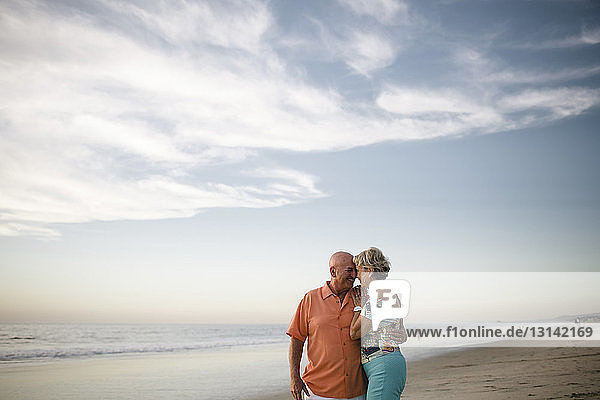 Romantisches älteres Ehepaar schaut sich an  während es bei Sonnenuntergang am Meer gegen den Himmel steht