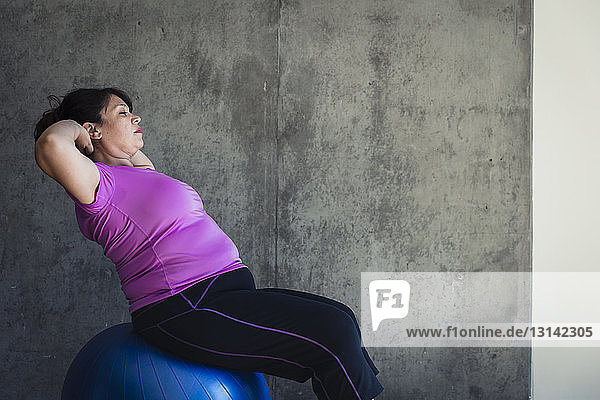Frau mit Händen hinter dem Kopf sitzend auf Fitnessball gegen Wand im Yogastudio