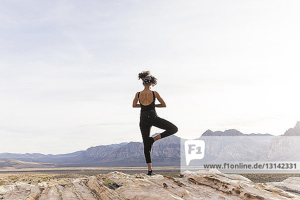 Rear view of woman meditating while standing on rock formation against sky