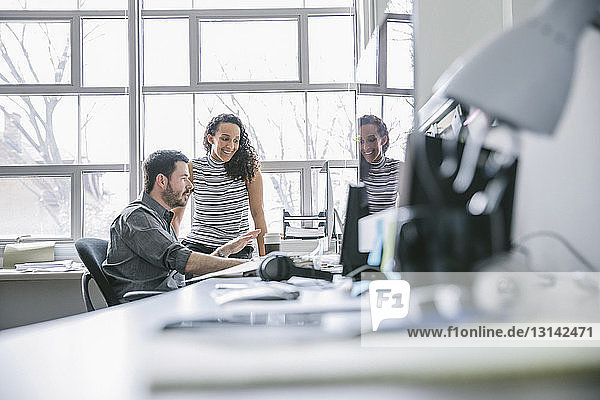 Lächelnde Geschäftsfrau steht einem männlichen Kollegen bei  der einen Desktop-Computer im Büro benutzt