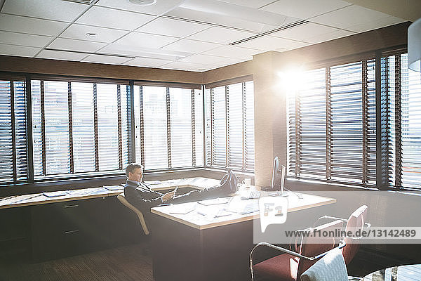 Businessman using mobile phone while relaxing in office during sunny day