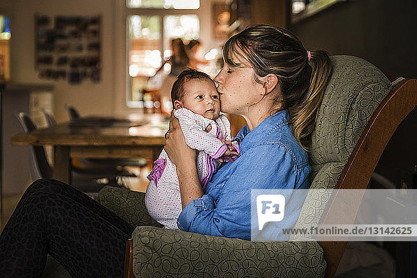 Seitenansicht einer Mutter  die ihre Tochter küsst  während sie zu Hause auf dem Sofa sitzt