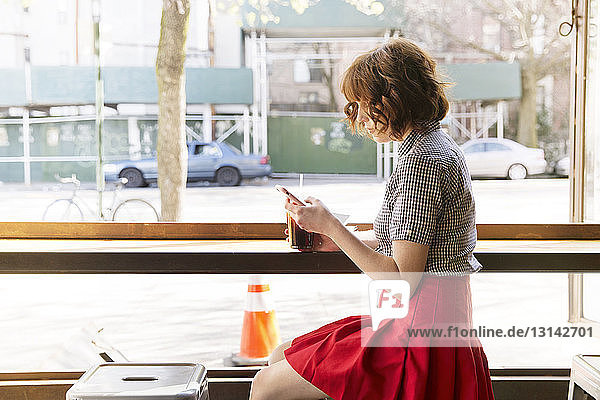 Seitenansicht einer Frau  die ein Smartphone benutzt  während sie im Cafe am Fenster sitzt