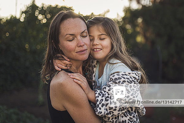 Mother and daughter with eyes closed embracing in yard during sunset