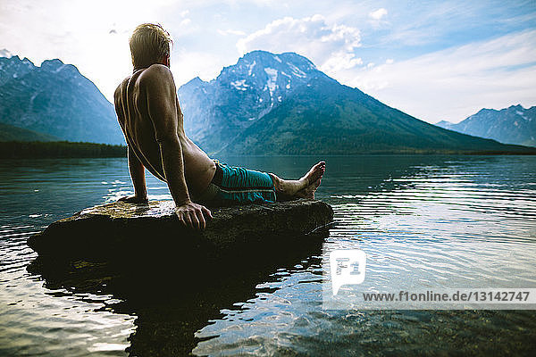Mann ohne Hemd schaut auf die Aussicht  während er sich auf einem Felsen im See gegen die Berge entspannt