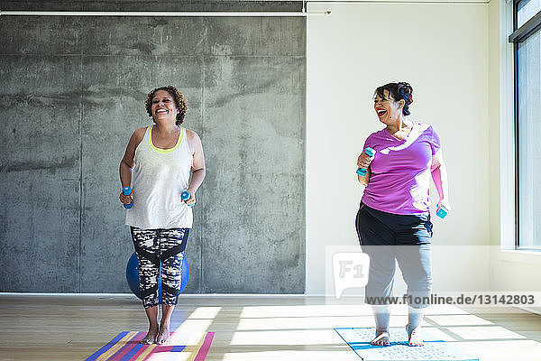 Full length of happy female friends using dumbbells while exercising against wall in yoga studio