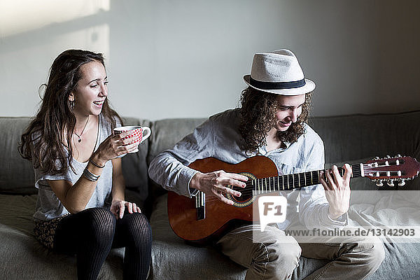 Glückliche Frau hält Kaffeetasse in der Hand und schaut dem Gitarre spielenden Bruder beim Sitzen auf dem Sofa zu