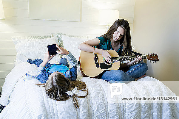 Girl using smart phone while sister playing guitar on bed at home