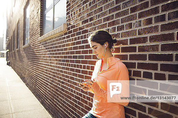 Sportliche Frau nutzt Smartphone an Ziegelsteinmauer an einem sonnigen Tag