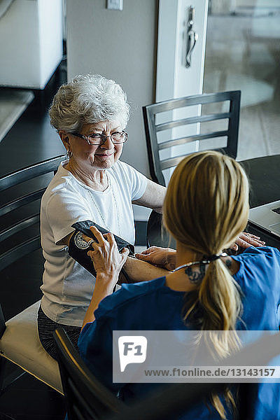 Schrägansicht der häuslichen Pflegekraft beim Einstellen des Blutdrucks Druckmesser am Arm einer älteren Frau am Esstisch