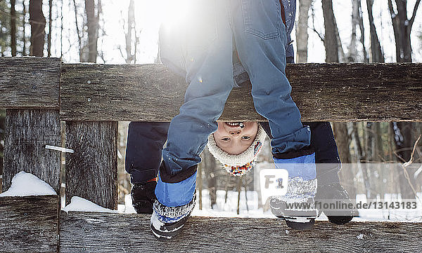 Portrait of happy boy bending while standing on fence during winter