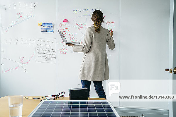 Rückansicht einer Geschäftsfrau  die auf einem Whiteboard schreibt  während sie im Büro über Sonnenkollektoren arbeitet