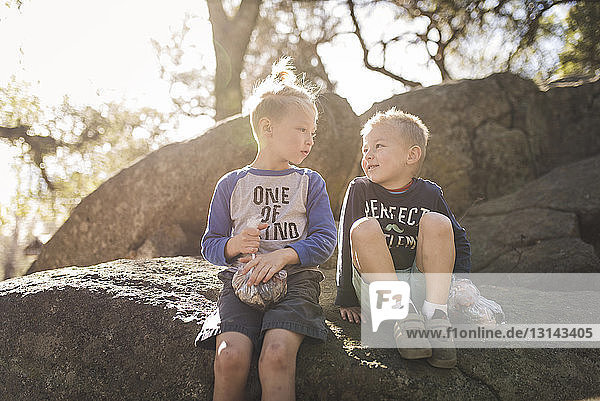 Brothers looking at each other while sitting on rocks against sky