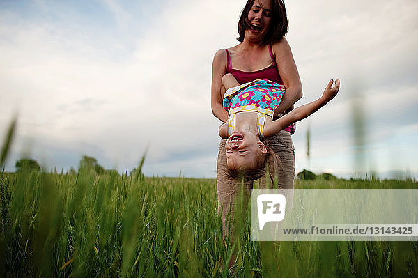 Low angle view of playful mother carrying daughter upside down on grassy field against cloudy sky