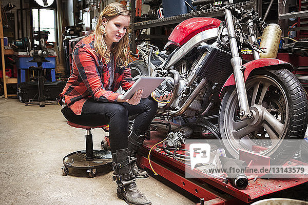 Frau benutzt digitales Tablett  während sie in der Werkstatt mit dem Fahrrad auf einem Hocker sitzt