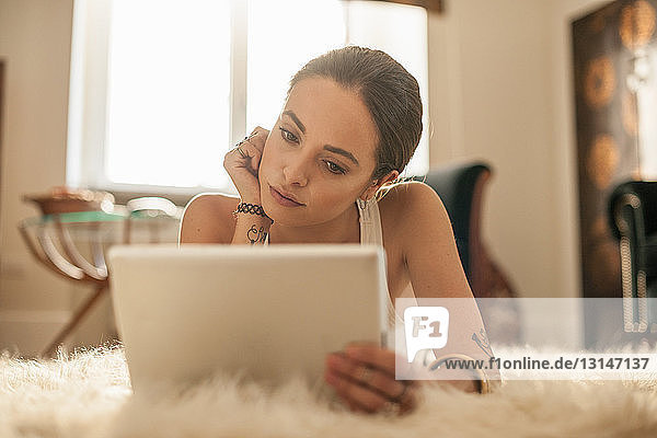 Junge Frau liegt auf einem Wohnzimmerteppich und schaut auf ein digitales Tablet