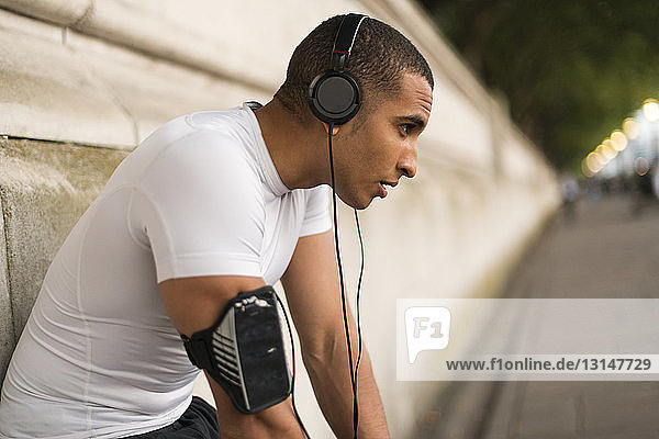 Erschöpfter männlicher Läufer mit Kopfhörern macht eine Pause am Flussufer