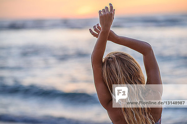 Rückansicht einer jungen Frau mit erhobenen Armen am Strand bei Sonnenuntergang