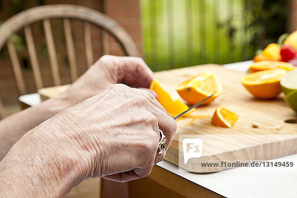 Hände einer älteren Frau beim Schneiden von Orangen am Gartentisch