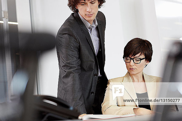 Businesswoman sitting at desk man looking over her shoulder at paperwork