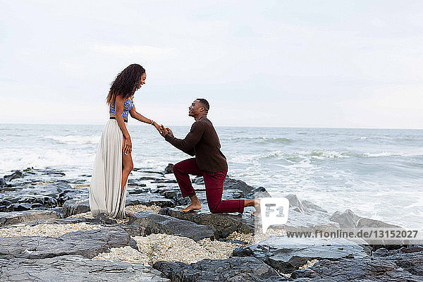 Mittlerer erwachsener Mann kniet auf Felsen am Meer und macht einer jungen Frau einen Heiratsantrag