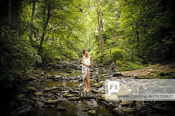 Mid adult woman standing barefoot on rocky riverbed squeezing water from hem of white dress