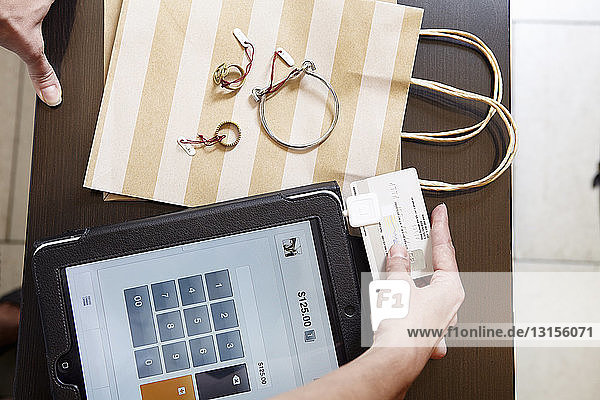 Mature woman using digital tablet to charge customer credit card in fashion boutique  focus on hands  overhead view