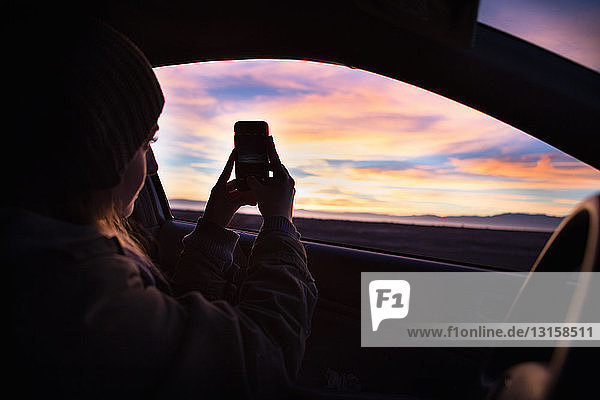 Junge Frau fotografiert den Sonnenuntergang mit ihrem Smartphone aus dem Auto heraus
