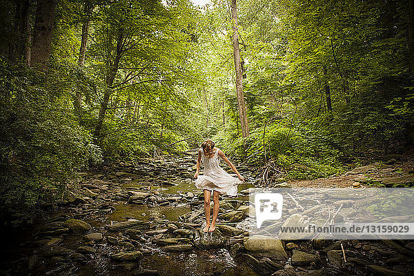 Mittlere erwachsene Frau steht barfuß auf einem felsigen Flussbett und wirbelt den Saum ihres weißen Kleides herum  während sie nach unten schaut