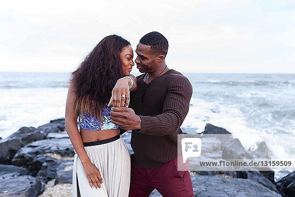 Paar steht auf Felsen am Meer und zeigt den Verlobungsring der Frau
