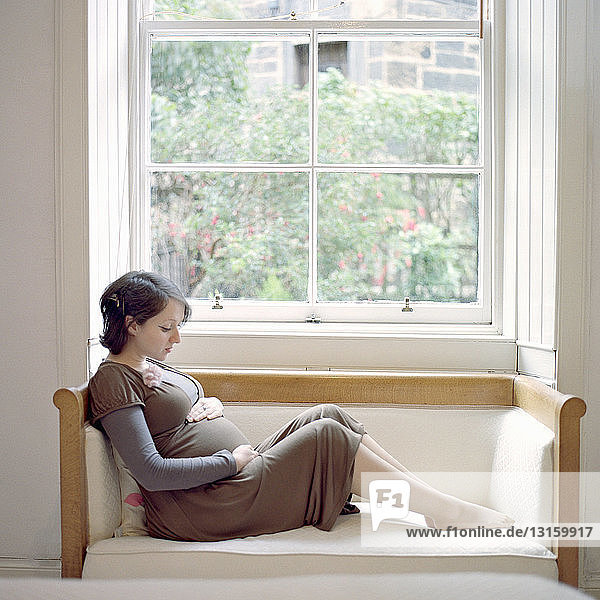 Porträt einer mittelgroßen schwangeren Frau  die auf einem Fensterplatz sitzt und den Bauch hält