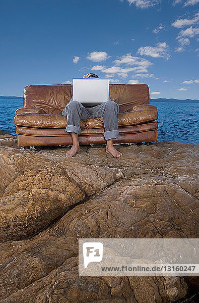 Mann auf Couch am Strand mit Laptop