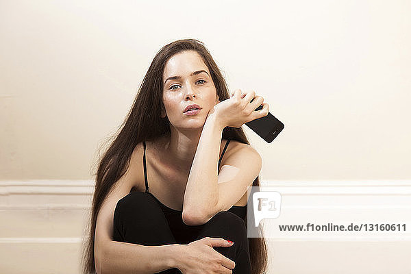 Young woman holding smartphone sitting hugging knees looking at camera