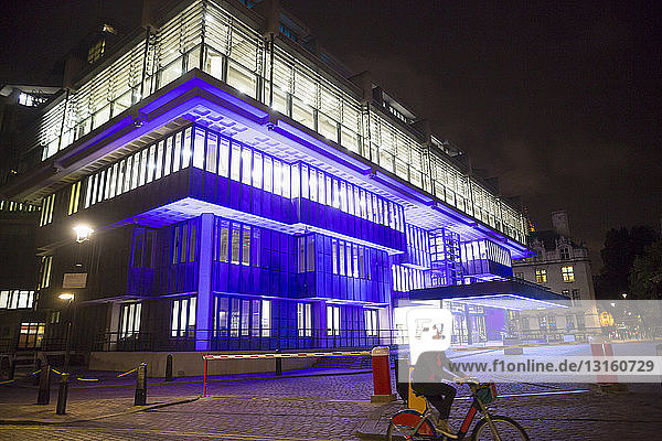 Ältere Frau radelt nachts an einem blau beleuchteten Bürogebäude vorbei  London  UK
