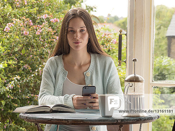 Porträt einer jungen Frau  die am Tisch sitzt und ein Smartphone hält