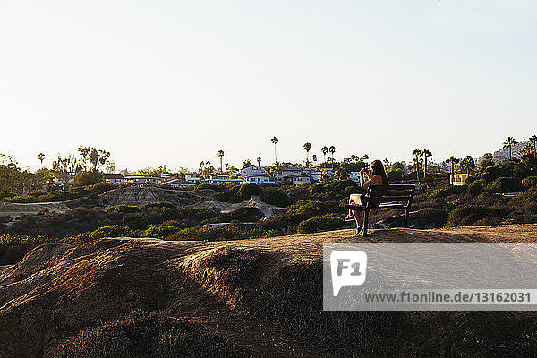 Rear view of young woman sitting on clifftop bench photographing view on smartphone  San Clemente  California  USA