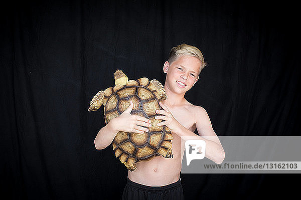 Vorderansicht eines Jungen mit nacktem Oberkörper  der eine afrikanische Spornschildkröte hält  in die Kamera blickend