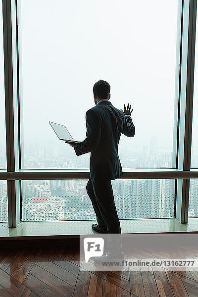 Junger Geschäftsmann mit Laptop  der aus dem Fenster eines Bürohochhauses nach unten blickt  Shanghai  China