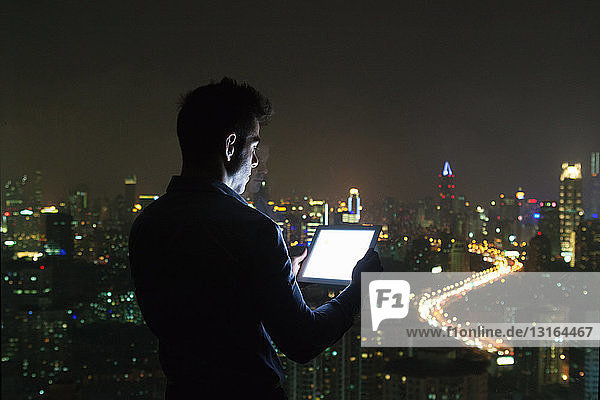 Silhouettierter junger Geschäftsmann betrachtet digitales Tablet vor dem Fenster eines Bürohochhauses bei Nacht  Shanghai  China