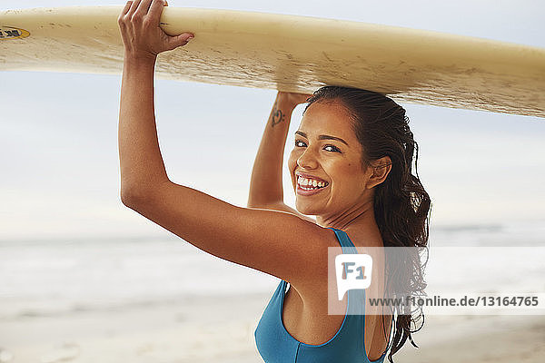 Porträt einer lächelnden jungen Frau mit einem Surfbrett auf dem Kopf am Strand  San Diego  Kalifornien  USA