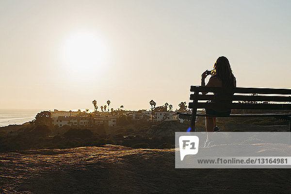 Silhouette of young woman sitting on clifftop bench photographing view on smartphone  San Clemente  California  USA