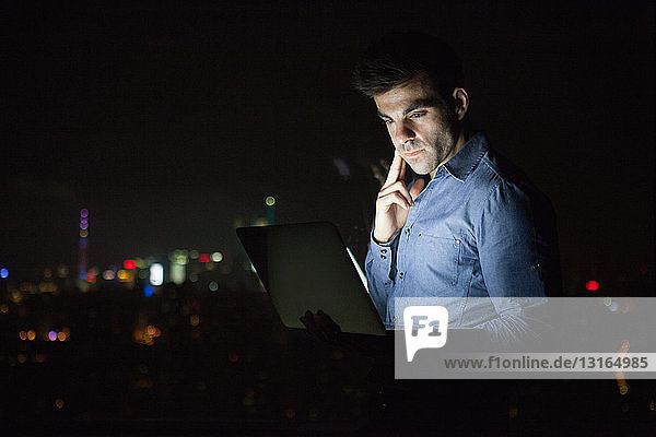 Young businessman looking at laptop in front of skyscraper office window at night  Shanghai  China
