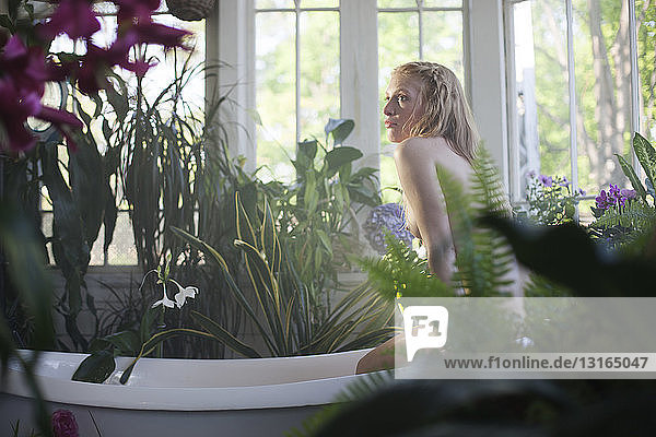 Nackte junge Frau sitzt auf dem Rand einer Badewanne  im Badezimmer voller Pflanzen
