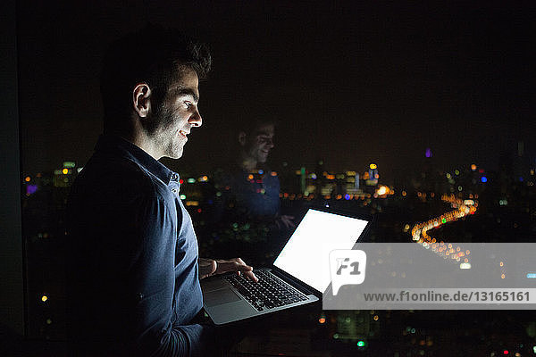 Young businessman typing on laptop in front of skyscraper office window at night  Shanghai  China
