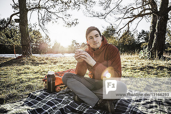 Young man sitting on picnic blanket drinking coffee in forest