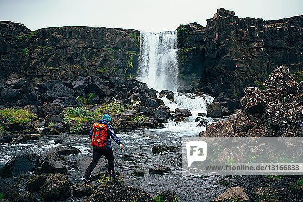 Rear view of mid adult woman with backpack stepping over rocks in river in front of waterfall  Iceland