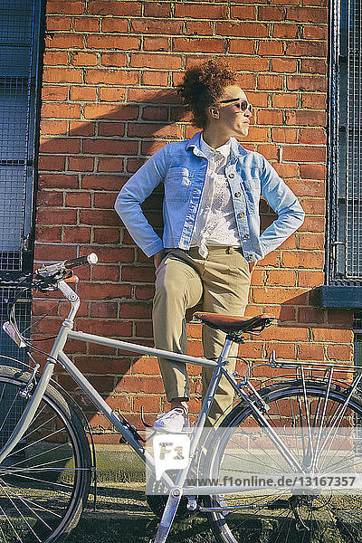 Junge Frau mit Fahrrad und Sonnenbrille lehnt mit den Händen auf der Hüfte an Ziegelmauer und schaut weg