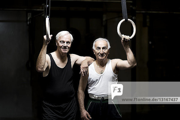 Portrait of two senior men holding gym rings in dark gym